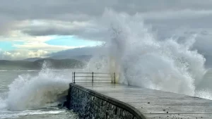 BBC Weather Watchers / snaphappydave Powerful waves crashing against a stone pier under a cloudy sky, showcasing the force of storms