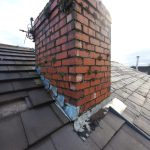 Chimney Repair. Close-up of a brick chimney with moss and dirt on a roof. The roof has dark gray tiles, and the sky is partly cloudy.