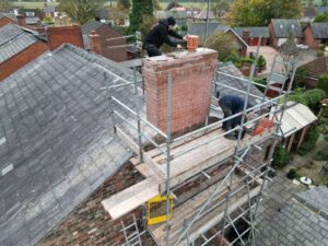 Two workers on scaffolding repairing a brick chimney on a house. One is kneeling, the other standing. The house has a gray tiled roof, and there are trees and other houses nearby.